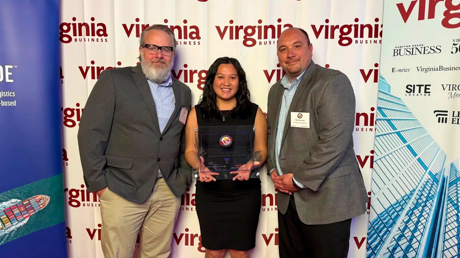Three employees standing in front of a Virginia Business media backdrop while holding a workplace award