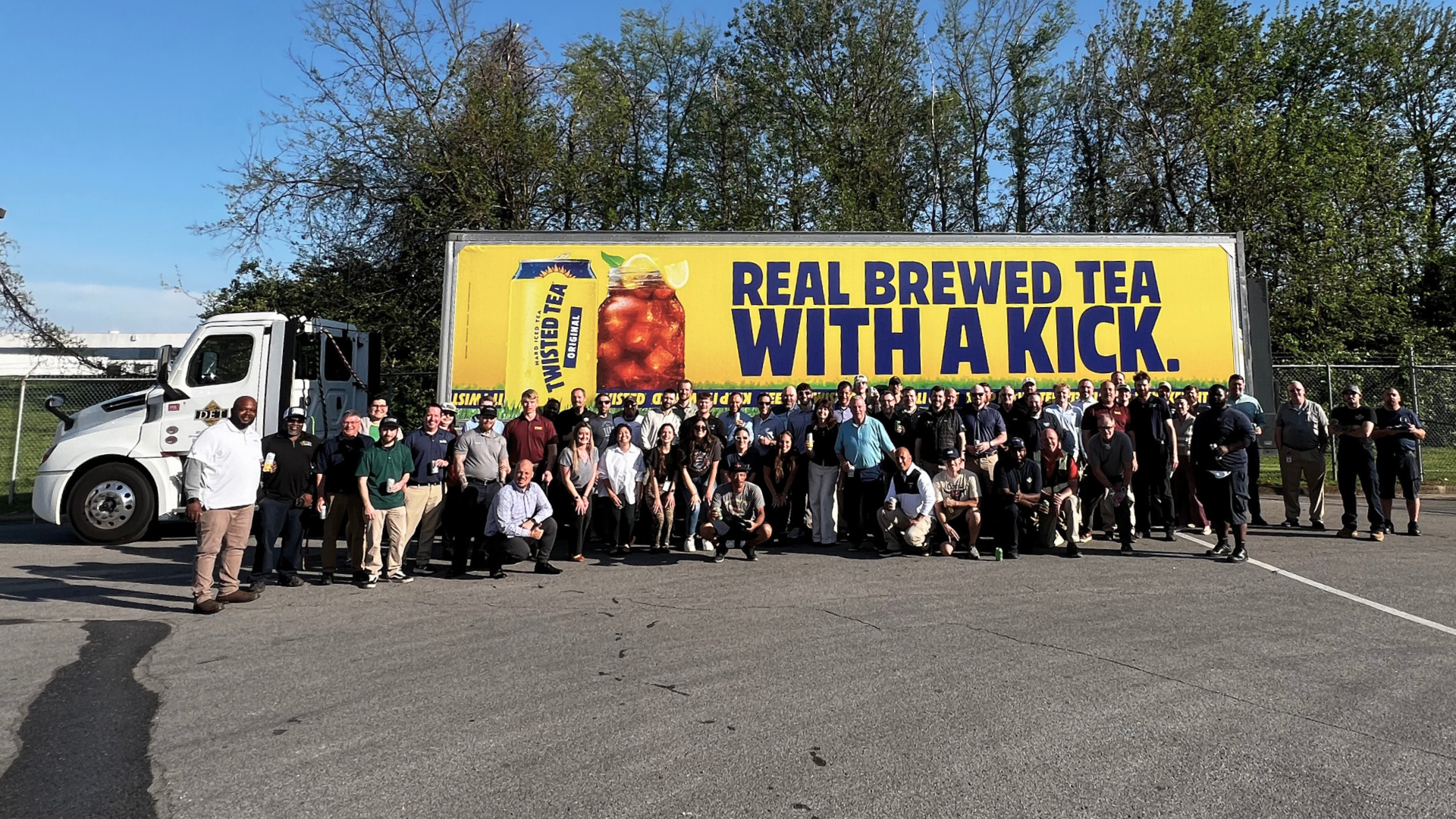 group of people smiling and posing in front of a delivery truck in a parking lot with a Twisted Tea truck wrap