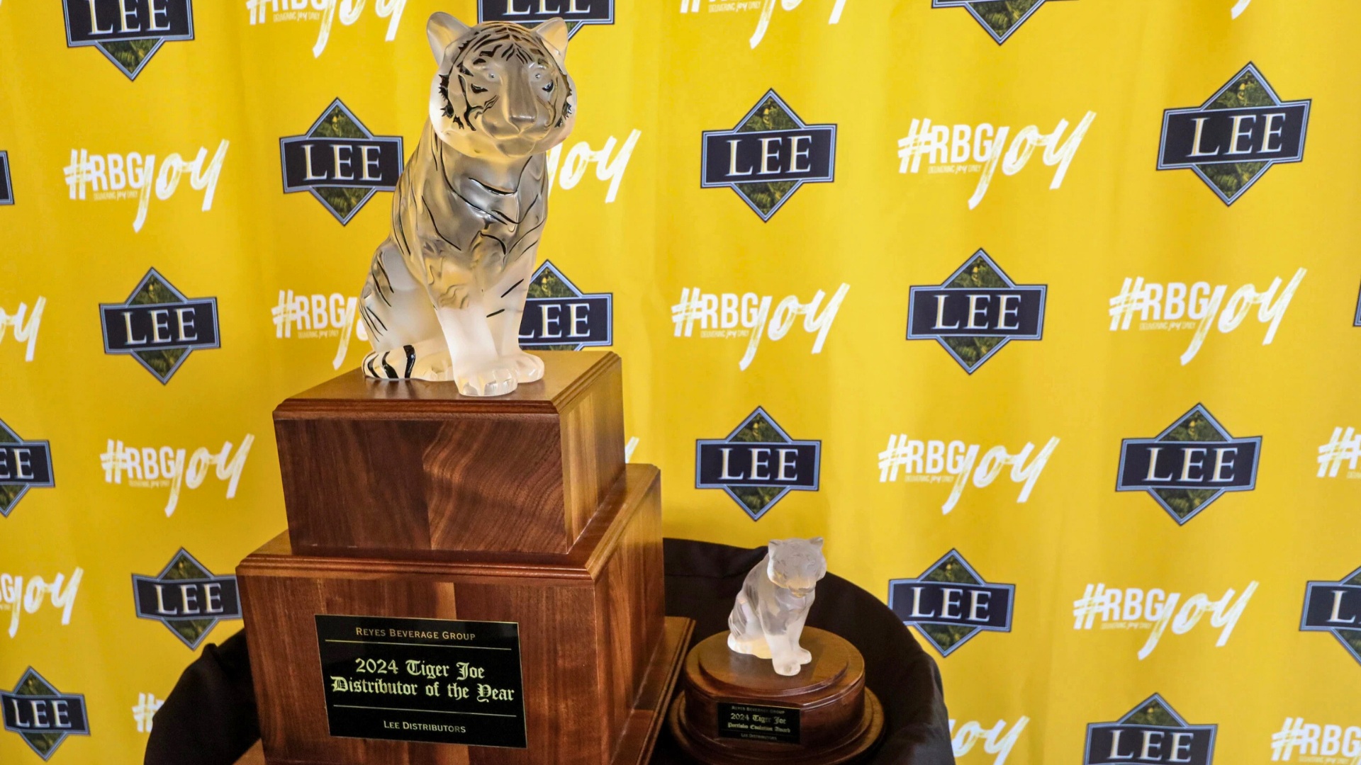 Large crystal Tiger Distributor of the Year award next to a smaller crystal tiger award on a table in front of a yellow backdrop with the #RBGJOY and Lee Distributors logo