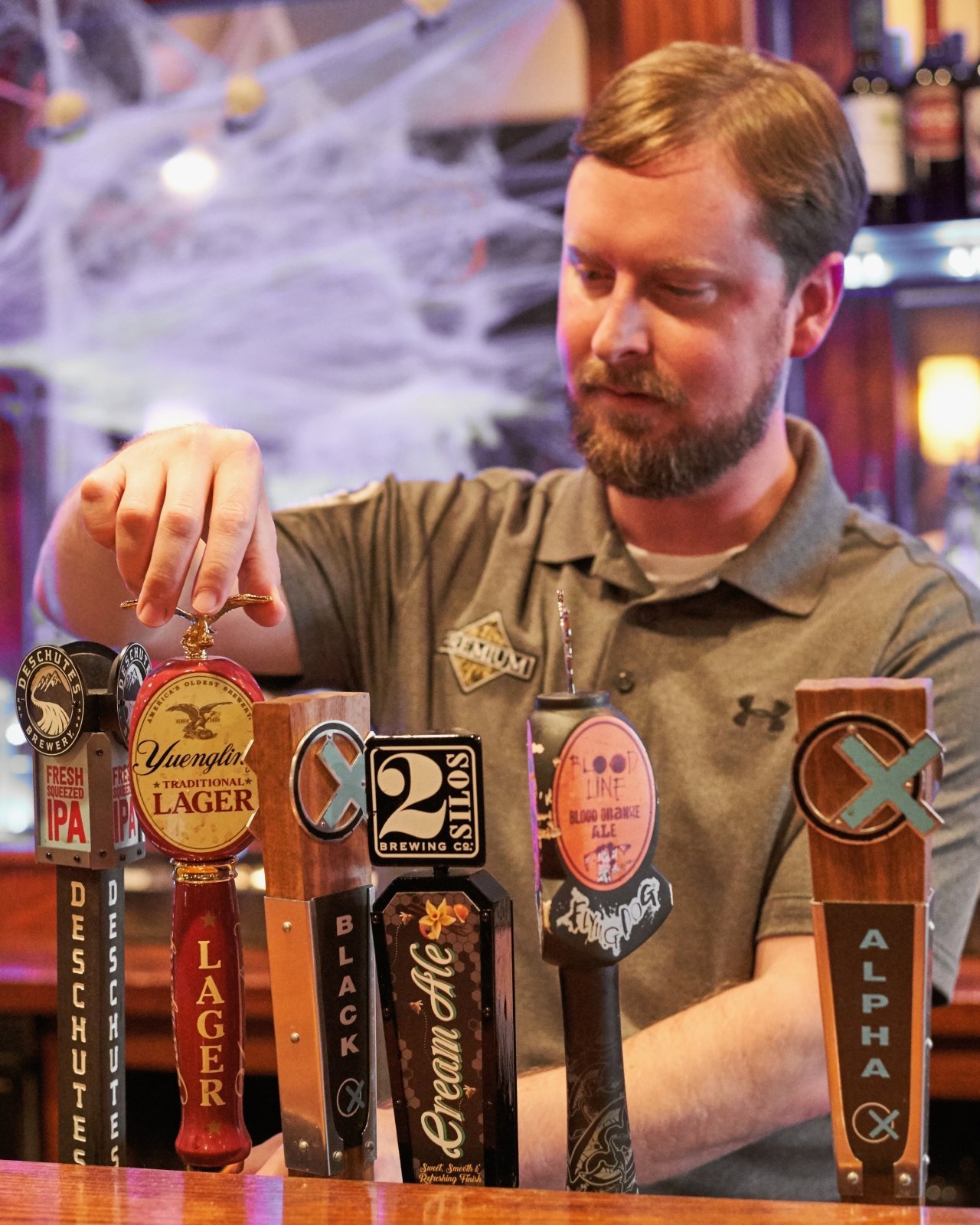 A man in a gray heathered polo type shirt holding the top of a tap handle in a bar setting