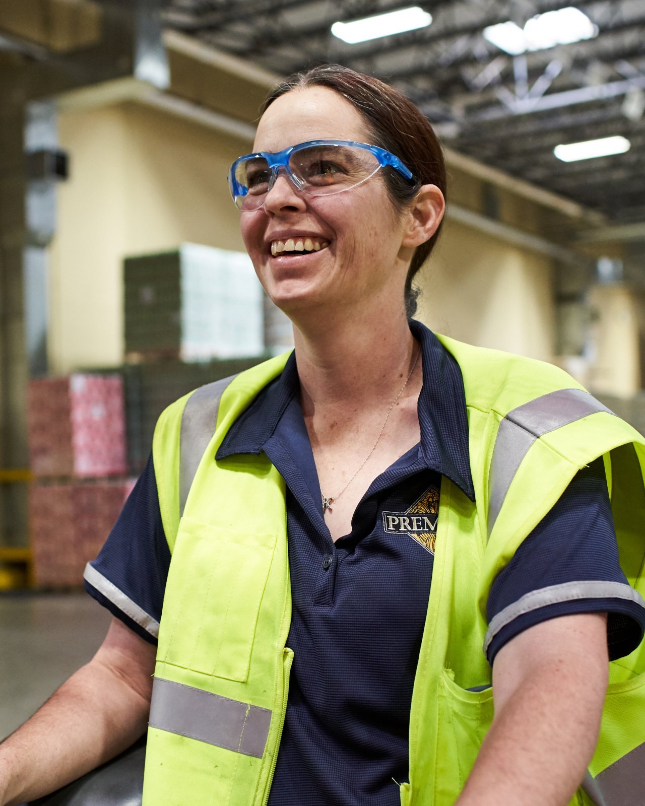 A woman with protective glasses, wearing a navy polo type shirt in a warehouse setting
