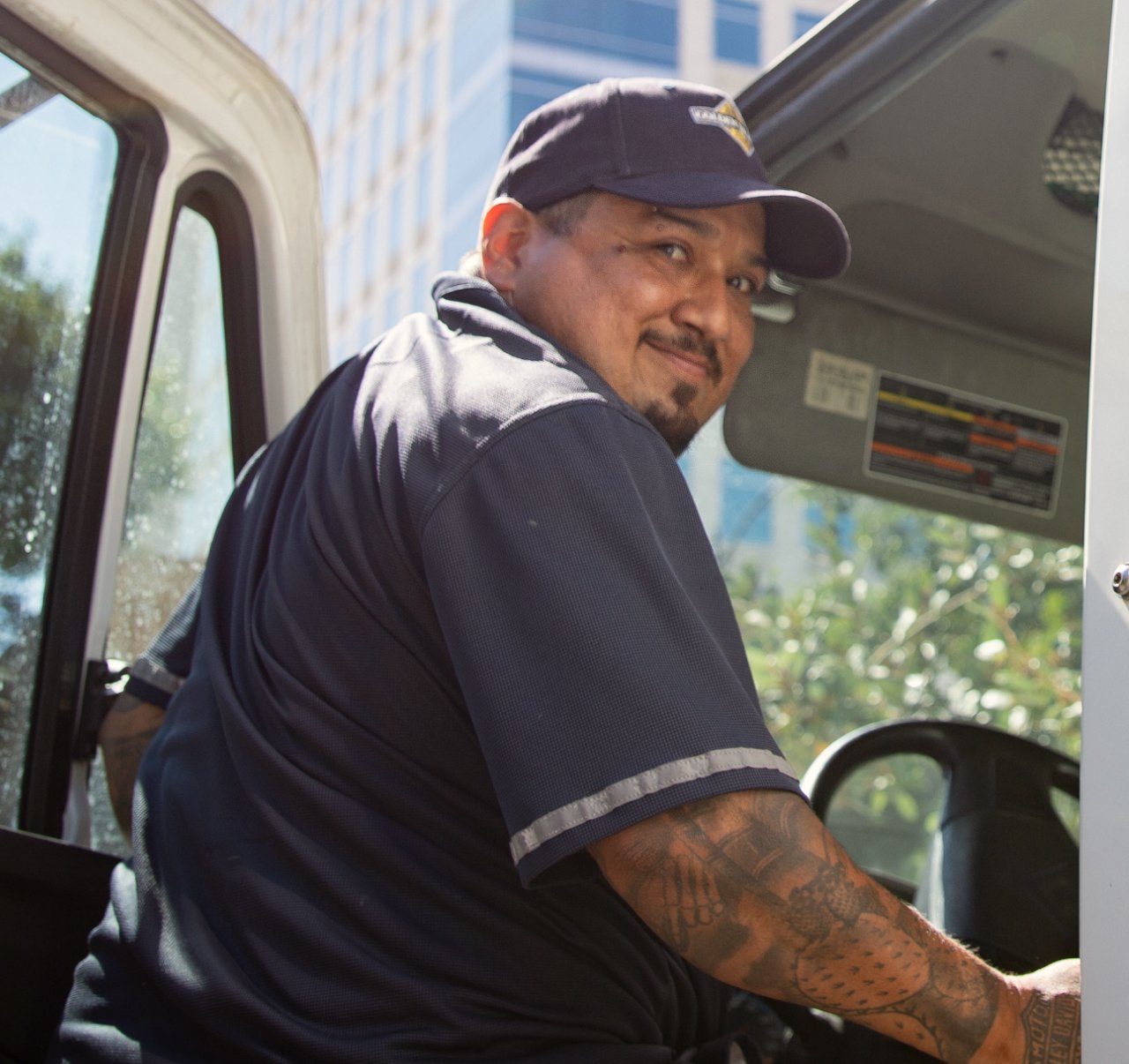 A man wearing a navy hat in a navy polo type shirt with grey reflective band at the bottom of the sleeves getting into a delivery truck cab