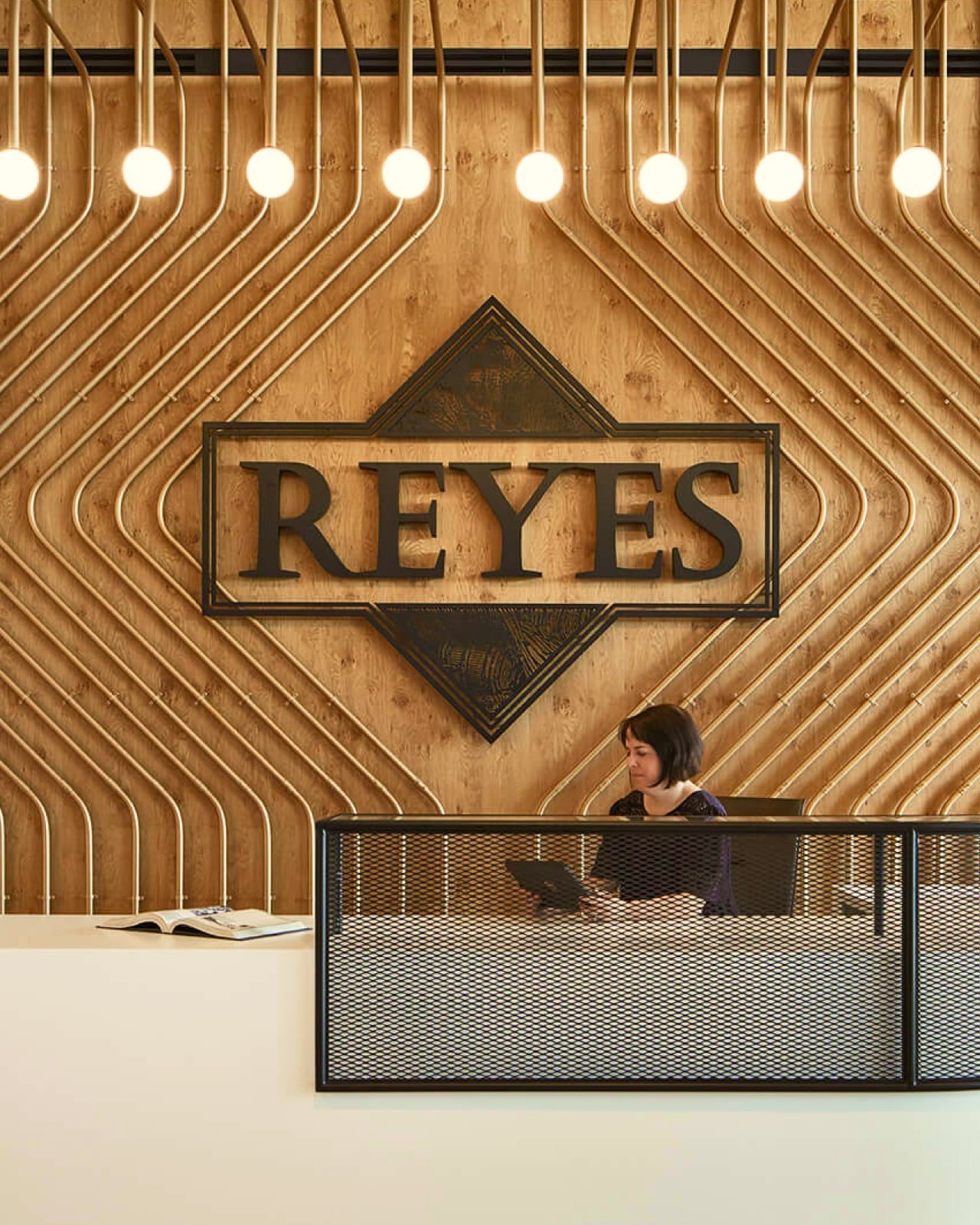 A wooden sign that shows the Reyes Beverage Group logo on a ornately designed light-colored wood wall with a woman sitting at a desk in front of it
