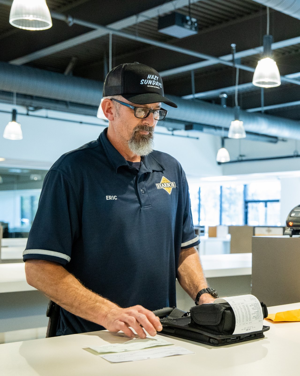 A man wearing glasses and a black hat and navy polo style shirt with a grey beard and black glasses standing inside the Harbor Distributing Delivery area printing off his orders