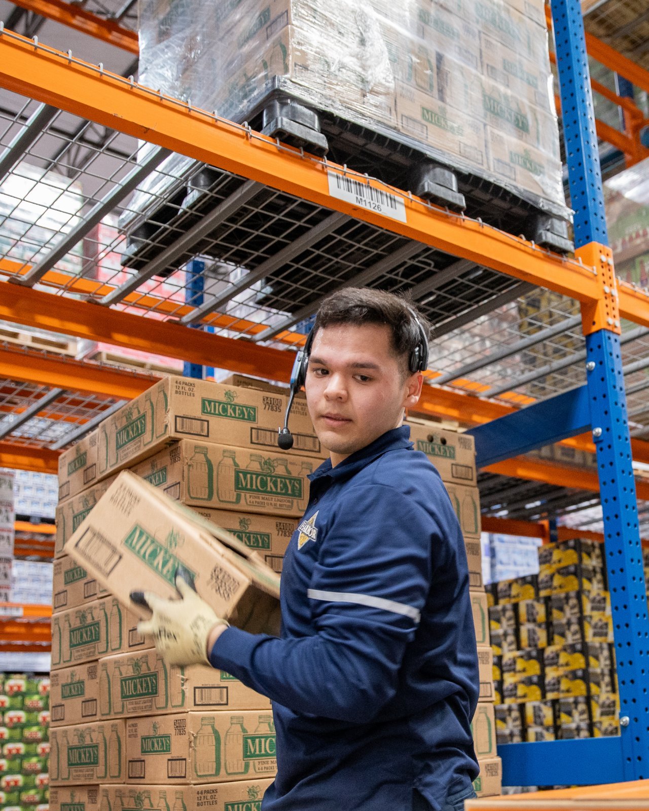 A man wearing a headset, a navy long sleeve shirt and yellow gloves lifting boxes in a warehouse